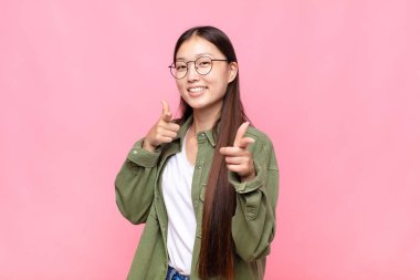 Asyalı genç bir kadın pozitif, başarılı, mutlu bir tavırla kamerayı işaret ederek, elleriyle silah işareti yapıyor.
