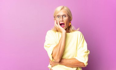Orta yaşlı bir kadın şok ve inançsızlık içinde, elleri yanağında ve kolları çaprazlanmış, sersemlemiş ve şaşırmış hissediyor.