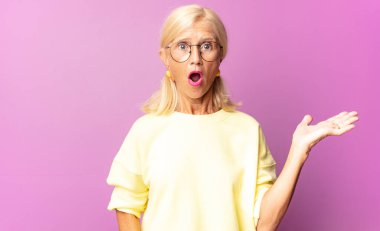 Orta yaşlı kadın şaşırmış ve şoka uğramış görünüyor, çenesi düşmüş ve eli açık bir nesneyi tutuyor.