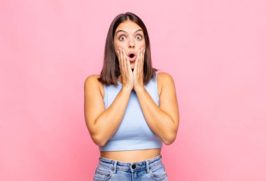 Genç ve güzel bir kadın şok olmuş ve korkmuş hissediyor, ağzı açık ve elleri yanaklarında korkmuş görünüyor.