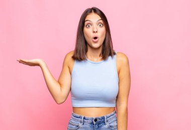 Genç ve güzel bir kadın şaşırmış ve şok olmuş görünüyor, çenesi düşmüş, bir objeyi elinde açık bir elle tutuyor.