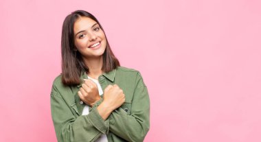 Genç, güzel bir kadın, neşeli bir şekilde gülümsüyor ve kutluyor. Yumruklarını sıkmış, kollarını kavuşturmuş, mutlu ve pozitif hissediyor.