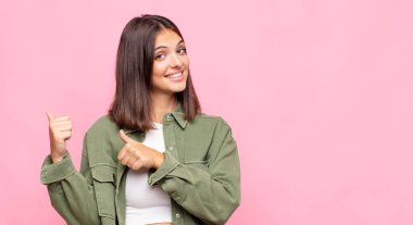 Genç ve güzel bir kadın neşeyle gülümsüyor ve yan taraftaki fotokopi alanını işaret ediyor, mutlu ve tatmin olmuş hissediyor.