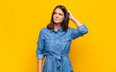 Genç, güzel bir kadın şaşkın ve kafası karışmış hissediyor, başını kaşıyor ve yan tarafa bakıyor.