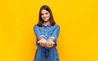 Genç, güzel bir kadın mutlu bir şekilde gülümsüyor. Dost canlısı, kendinden emin, olumlu bakışlı, bir nesne ya da konsept sunuyor ve gösteriyor.