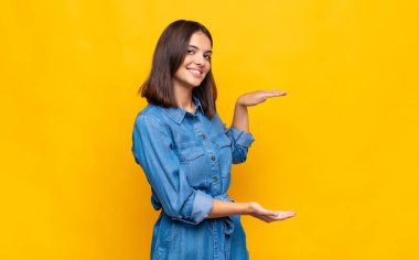 Genç, güzel bir kadın gülümsüyor, mutlu, pozitif ve tatmin olmuş hissediyor, fotokopi alanında nesne veya konsept tutuyor veya gösteriyor