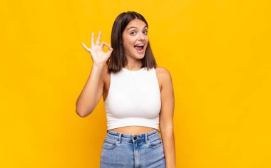 Genç ve güzel bir kadın kendini başarılı ve tatmin olmuş hissediyor, ağzı açık gülümsüyor, elle işaret veriyor.