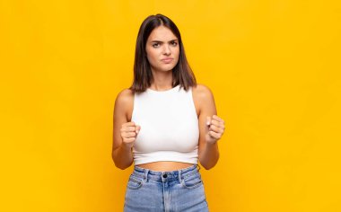 Genç ve güzel bir kadın kendinden emin, sinirli, güçlü ve agresif görünüyor. Yumrukları boks pozisyonunda dövüşmeye hazır.