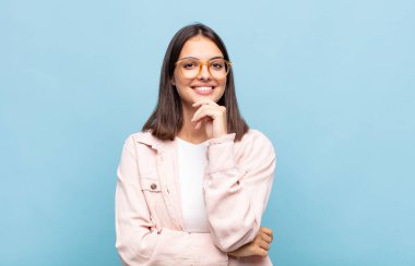 Genç ve güzel bir kadın mutlu görünüyor ve gülümsüyor. Elleri çenesinde, merak ediyor ya da soru soruyor. Seçenekleri karşılaştırıyor.