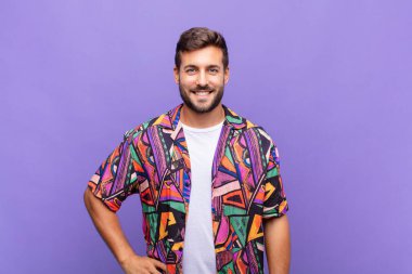 Genç adam mutlu bir şekilde gülümsüyor, eli kalçasında ve kendinden emin, olumlu, gururlu ve arkadaş canlısı.