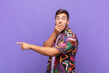 Genç adam mutlu, şaşkın ve şaşırmış hissediyor. Ağzını eliyle kapatıyor ve yanal fotokopi alanını işaret ediyor.