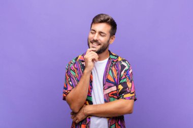 Genç adam mutlu, kendinden emin bir ifadeyle gülümsüyor, çenesinde elleri olan, merak eden ve yan tarafa bakan