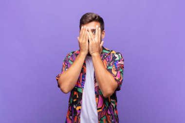 Genç adam elleriyle yüzünü kapatıyor, parmaklarının arasından şaşkın bir ifadeyle yan tarafa bakıyor.