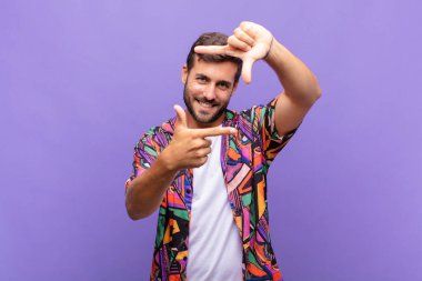 Genç adam mutlu, arkadaş canlısı ve pozitif hissediyor, gülümsüyor ve elleriyle bir portre veya fotoğraf çerçevesi yapıyor.