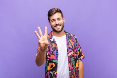 Genç adam gülümsüyor ve arkadaş canlısı görünüyor, bir numarayı gösteriyor ya da ilk eli önde, geriye doğru sayıyor.