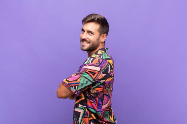 Genç adam neşeyle gülümsüyor, mutlu, tatmin olmuş ve rahatlamış hissediyor, kollarını kavuşturup yan tarafa bakıyor.