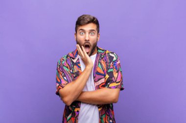 Genç adam şok ve inançsızlık içinde, elleri yanak ve kolları çapraz, sersemlemiş ve şaşırmış hissediyor.