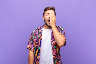 Genç adam uykulu, sıkılmış ve esniyor, baş ağrısı ve yüzünün yarısını kaplayan bir elle.