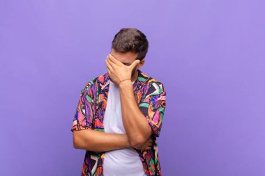 Genç adam stresli, utanmış ya da üzgün görünüyor, baş ağrısı var, yüzü elle kapatılmış.