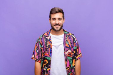 Genç adam pozitif ve kendinden emin bir şekilde gülümsüyor, tatmin olmuş, arkadaş canlısı ve mutlu görünüyor.