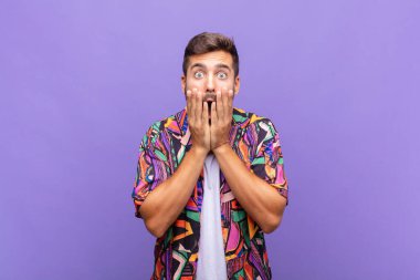 Genç adam şok olmuş ve korkmuş hissediyor, ağzı açık ve elleri yanaklarında korkmuş görünüyor.