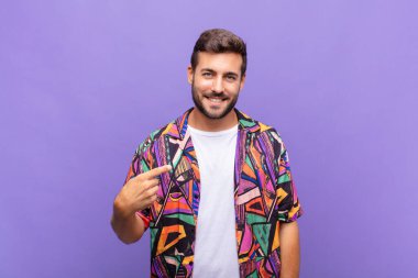 Genç adam gururlu, kendinden emin ve mutlu görünüyor, gülümsüyor ve kendini işaret ediyor veya bir numaralı işareti yapıyor.