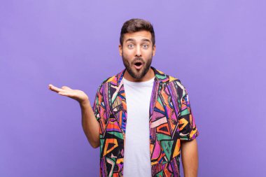 Genç adam şaşırmış ve şok olmuş görünüyor, çenesi düşmüş bir nesneyi elinde açık bir elle tutuyor.