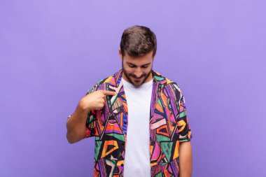 Genç adam neşeyle gülümsüyor, yere bakıyor ve göğsüne işaret ediyor.