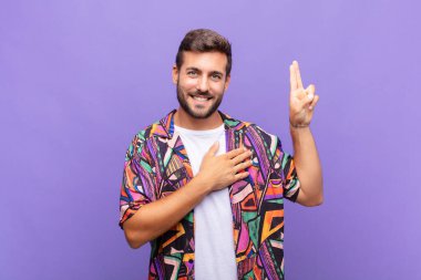 Genç adam mutlu, kendinden emin ve güvenilir görünüyor, gülümsüyor ve olumlu bir tavırla zafer işareti gösteriyor.