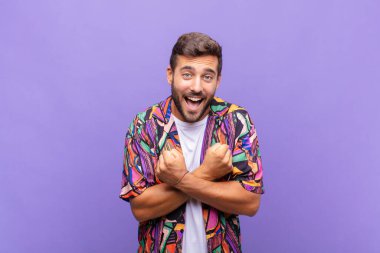 Genç adam sevinçle gülümsüyor ve kutluyor, yumruklarını sıkmış ve kollarını kavuşturmuş, mutlu ve pozitif hissediyor.