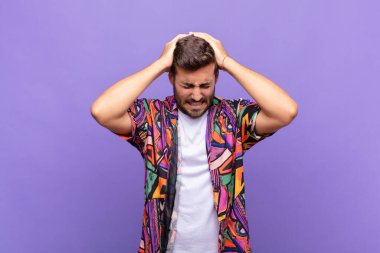 Genç adam kendini stresli ve hayal kırıklığına uğramış hissediyor, ellerinizi havaya kaldırıyor, yorgun, mutsuz ve migren ağrısı çekiyor.