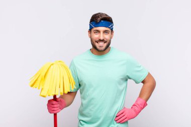 Genç adam mutlu bir şekilde gülümsüyor, eli kalçasında ve kendinden emin, olumlu, gururlu ve arkadaş canlısı.