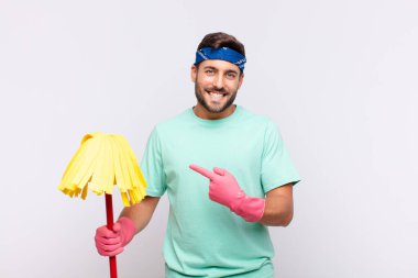 Genç adam neşeyle gülümsüyor, mutlu hissediyor ve kenarı ve yukarıyı işaret ediyor, fotokopi uzayındaki nesneleri gösteriyor.