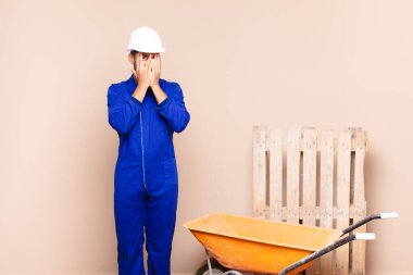 Genç adam elleriyle yüzünü kapatıyor, parmaklarının arasından şaşkın bir ifadeyle bakıyor ve yan inşaat konseptine bakıyor.