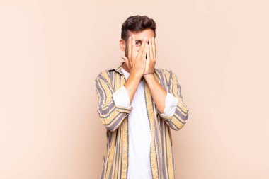 Genç adam elleriyle yüzünü kapatıyor, parmaklarının arasından şaşkın bir ifadeyle yan tarafa bakıyor.