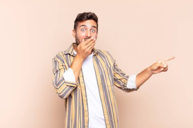 Genç adam mutlu, şaşkın ve şaşırmış hissediyor. Ağzını eliyle kapatıyor ve yanal fotokopi alanını işaret ediyor.