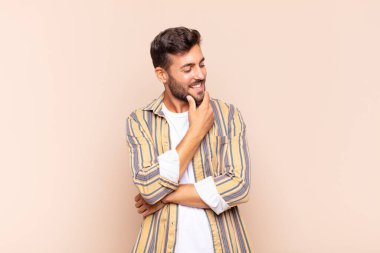 Genç adam mutlu, kendinden emin bir ifadeyle gülümsüyor, çenesinde elleri olan, merak eden ve yan tarafa bakan