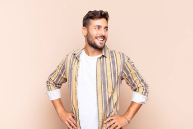 Genç adam mutlu, neşeli ve kendinden emin görünüyor, gururla gülümsüyor ve iki elini de kalçasında istiyor.