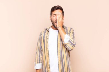 Genç adam uykulu, sıkılmış ve esniyor, baş ağrısı ve yüzünün yarısını kaplayan bir elle.