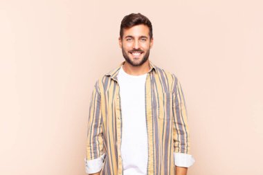 Genç adam pozitif ve kendinden emin bir şekilde gülümsüyor, tatmin olmuş, arkadaş canlısı ve mutlu görünüyor.