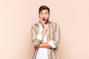 Genç adam şok ve inançsızlık içinde, elleri yanak ve kolları çapraz, sersemlemiş ve şaşırmış hissediyor.