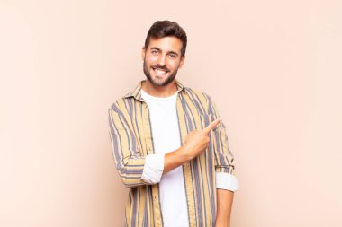 Genç adam neşeyle gülümsüyor, mutlu hissediyor ve kenarı ve yukarıyı işaret ediyor, fotokopi uzayındaki nesneleri gösteriyor.