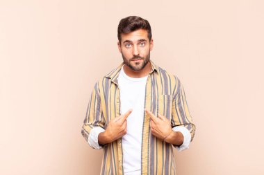 Genç adam şaşkın ve şaşkın bir bakışla kendini işaret ediyor. Seçildiğine şaşırmış ve şaşırmış.