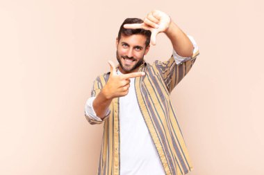 Genç adam mutlu, arkadaş canlısı ve pozitif hissediyor, gülümsüyor ve elleriyle bir portre veya fotoğraf çerçevesi yapıyor.