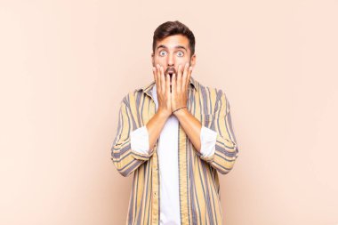 Genç adam şok olmuş ve korkmuş hissediyor, ağzı açık ve elleri yanaklarında korkmuş görünüyor.