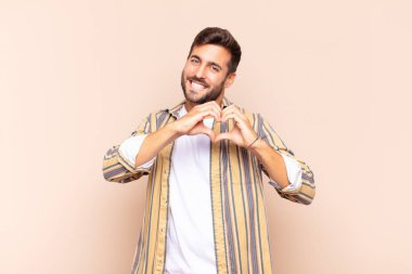 Genç adam gülümsüyor ve mutlu, sevimli, romantik ve aşık, iki eliyle kalbi şekillendiriyor.