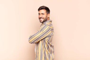 Genç adam neşeyle gülümsüyor, mutlu, tatmin olmuş ve rahatlamış hissediyor, kollarını kavuşturup yan tarafa bakıyor.