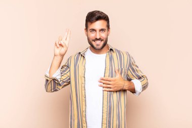 Genç adam mutlu, kendinden emin ve güvenilir görünüyor, gülümsüyor ve olumlu bir tavırla zafer işareti gösteriyor.