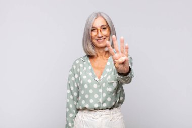 Gri saçlı kadın gülümsüyor ve arkadaş canlısı görünüyor. Elleri önde, geriye doğru sayıyor.