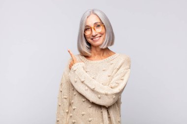 Gri saçlı kadın neşeyle gülümsüyor, mutlu hissediyor ve yan ve yukarı doğru işaret ediyor, fotokopi uzayındaki nesneleri gösteriyor